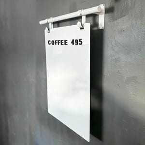 May include: White sign with black text that reads "COFFEE 495" hanging from a white metal rod attached to a gray wall.