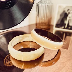 May include: Two vintage bangles with a cream and brown color scheme are displayed on a reflective surface. The bangles are stacked, showcasing their contrasting hues. A vintage photograph and a decorative glass bottle are in the background, along with a vinyl record.