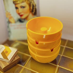 May include: Four bright yellow ashtrays stacked, each with a cigarette rest and a central post. A pack of Merit cigarettes and a gold cigarette case are also present. A vintage poster is in the background.