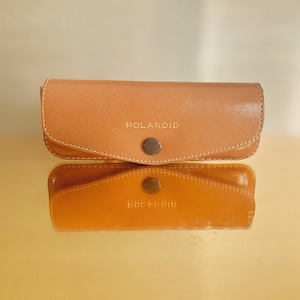 Puede incluir: Una funda de cuero marrón con la palabra "POLAROID" grabada en la parte delantera.