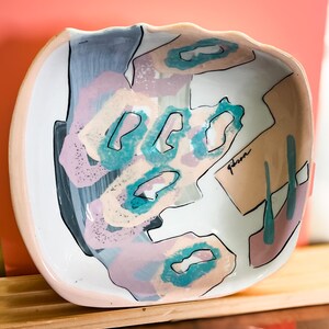 May include: A white ceramic bowl with a wavy rim. The bowl is decorated with abstract shapes in shades of pink, blue, and green. The word "glam" is written in black on the bowl.