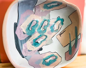 Może przedstawiać: Biała ceramiczna miska z falistym brzegiem. Miska jest ozdobiona abstrakcyjnymi kształtami w odcieniach różowego, niebieskiego i zielonego. Słowo "glam" jest napisane czarnym kolorem na misce.
