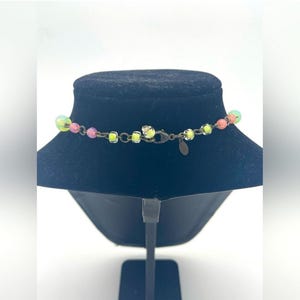 Op de afbeelding: Een kleurrijke ketting met een verscheidenheid aan kralen in roze, geel en groen. De ketting wordt getoond op een zwarte mannequin buste. De sluiting en ketting zijn donker bronskleurig.