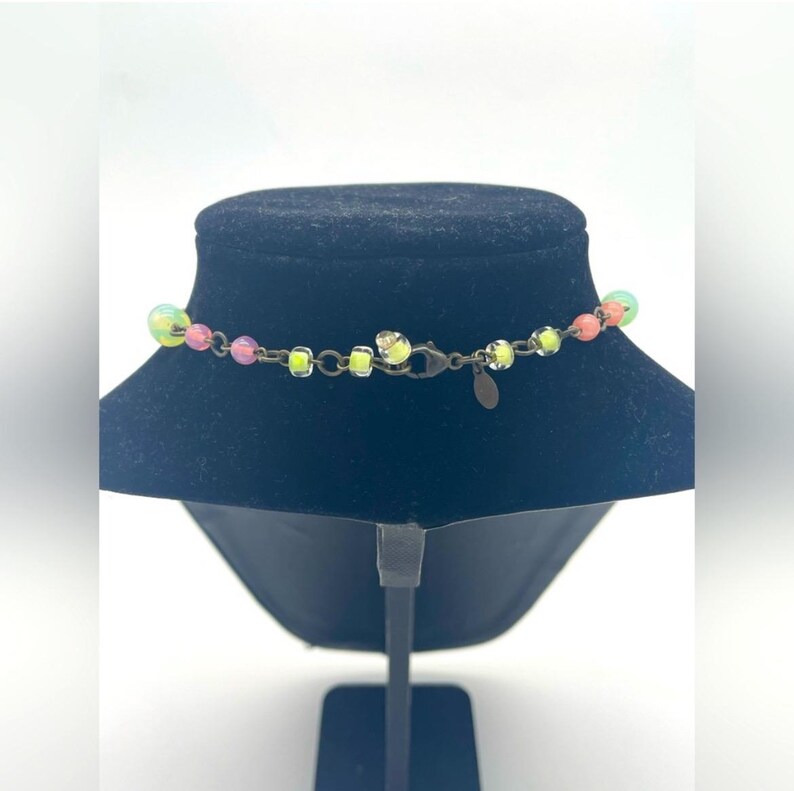 Op de afbeelding: Een kleurrijke ketting met een verscheidenheid aan kralen in roze, geel en groen. De ketting wordt getoond op een zwarte mannequin buste. De sluiting en ketting zijn donker bronskleurig.