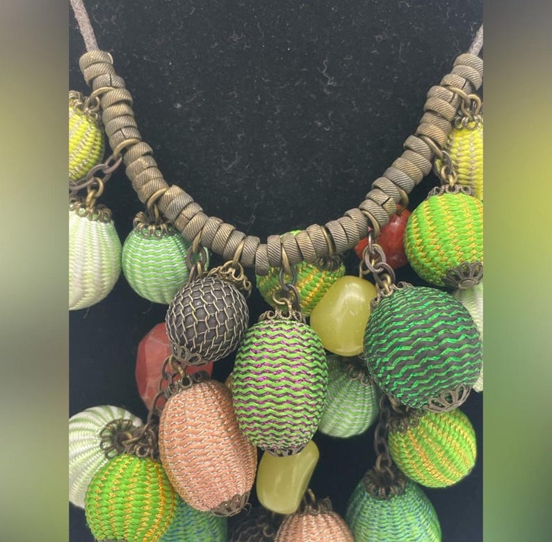 Op de afbeelding: Een kleurrijke ketting met diverse decoratieve elementen. De ketting heeft een bruin koord en een reeks hangende bedels in tinten groen, geel, oranje en rood. De bedels hebben een geweven textuur en metalen details.