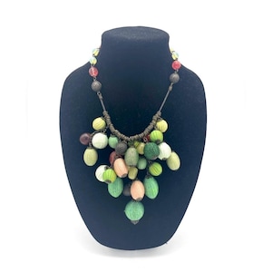 Op de afbeelding: Een kleurrijke kralenketting tentoongesteld op een zwarte buste. De ketting heeft een verscheidenheid aan groene, gele, roze en bruine kralen. De kralen zijn gegroepeerd, wat een uniek en opvallend ontwerp cre&euml;ert.