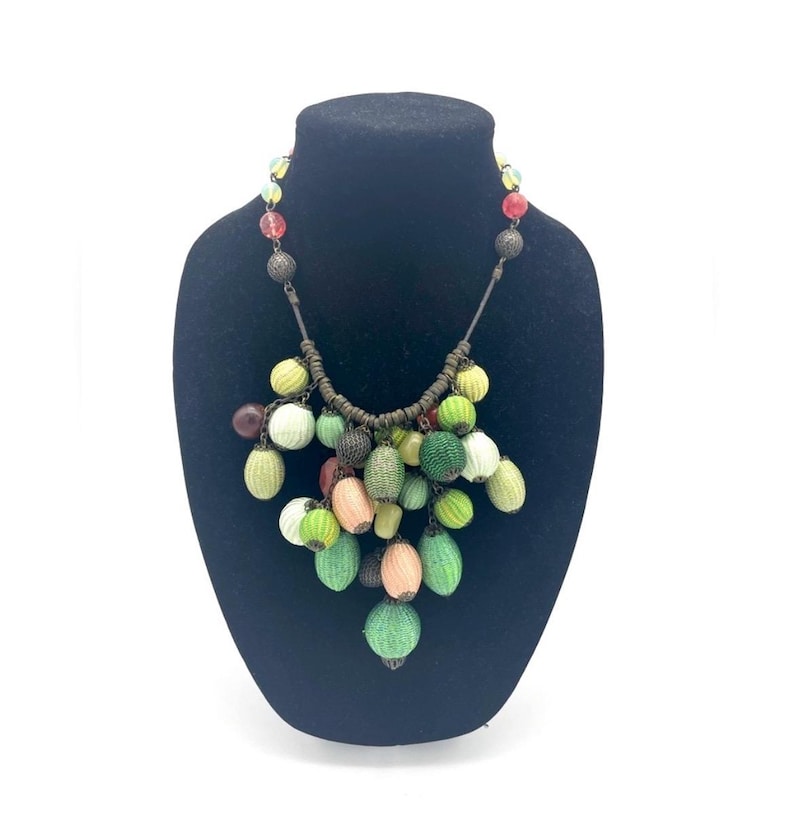 Op de afbeelding: Een kleurrijke kralenketting tentoongesteld op een zwarte buste. De ketting heeft een verscheidenheid aan groene, gele, roze en bruine kralen. De kralen zijn gegroepeerd, wat een uniek en opvallend ontwerp cre&euml;ert.