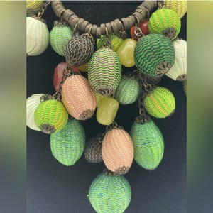 Op de afbeelding: Een kleurrijke ketting met diverse geweven en met kralen bezette bedels in groen, roze en geel. De ketting heeft een donkerbruin koord en metalen kettingaccenten. De bedels hebben de vorm van bollen en ovalen.