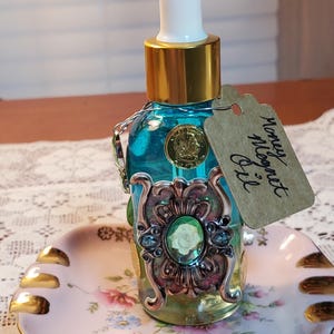May include: A small glass bottle with a gold top and a dropper. The bottle is filled with a blue liquid and decorated with a metal embellishment and a green gemstone. The bottle is sitting on a pink and white porcelain dish with a gold rim. A brown paper tag is attached to the bottle with the text "Honey Moon Oil".