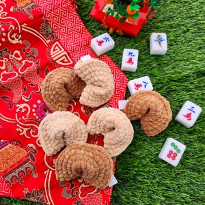 May include: A group of six brown and beige crocheted fortune cookies on a red and gold patterned fabric. The fabric is on top of green grass. There are also several white tiles with Chinese characters on them.