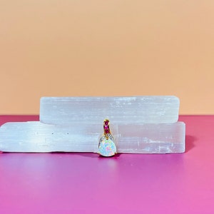 Może przedstawiać: Wisiorek z perłowego białego opalu z złotą oprawą i rzędem różowych kamieni szlachetnych. Wisiorek leży na białym krysztale selenitu.