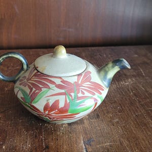 Op de afbeelding: Een keramische theepot met een crèmekleurige body, versierd met rode bloempatronen en groene bladeren. Het deksel en de handgreep zijn crèmekleurig en de tuit is donkergroen. De theepot staat op een houten oppervlak.