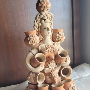 May include: A terracotta ceramic sculpture of a woman adorned with floral details and holding multiple small vases. The sculpture features a detailed face, braided hair, and is decorated with flower-shaped accents. The color palette is primarily earthy tones.