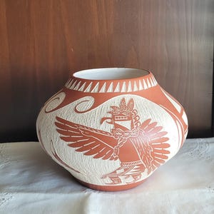 Könnte beinhalten: Ein terrakottafarbener Keramiktopf mit einem breiten, bauchigen Körper und einer schmalen Öffnung. Die Außenseite ist mit aufwendigen, geschnitzten weißen Mustern verziert, die eine stilisierte Vogelfigur mit ausgebreiteten Flügeln und geometrische Motive zeigen.