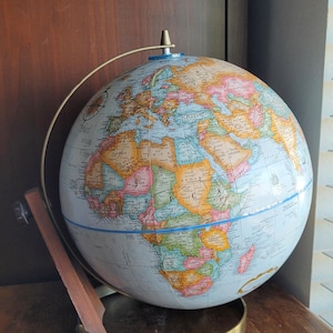 Peut inclure: Un globe vintage avec une carte du monde détaillée, représentant les pays en différentes couleurs. Il est monté sur un support en métal couleur laiton avec un bras de support en bois. Le globe mesure environ 30 cm de diamètre.