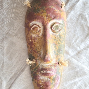 Könnte beinhalten: Eine längliche, ovale Wandmaske mit einer gesprenkelten, abgenutzten Lackierung in den Farben Dunkelrot, Olivgrün und Senfgelb. Die Maske hat geschnitzte, mandelförmige Augen und Lippen, die weiß hervorgehoben sind, sowie vier kleine Seilbindungen.