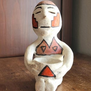 May include: A handcrafted ceramic figurine of a seated person with closed eyes. The figure is painted in white with orange and black geometric designs on the face and body. It holds a small bowl, and the overall style suggests an ancient or tribal aesthetic.