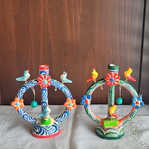 May include: Two circular ceramic candle holders featuring hand-painted floral and swirl patterns. The left holder is blue and white with light blue birds, while the right holder is green and white with yellow birds. Both have central flower motifs.