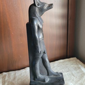 May include: A black statue of the Egyptian deity Anubis, depicted with a jackal head and human body. The figure stands in a profile view, wearing a traditional kilt and a striped headdress, attached to a vertical rectangular back support.