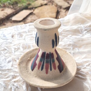 May include: A small, off-white ceramic candle holder with a matching saucer. The candle holder has a cylindrical shape with blue and maroon painted accents. The saucer is round and has a textured surface. The candle holder is sitting on a white lace tablecloth.