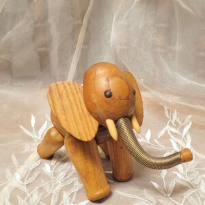 May include: Wooden elephant figurine with a light brown finish. The elephant has a rounded head, large ears, and a coiled metal trunk. The figurine is approximately 13 cm long and 10 cm tall.