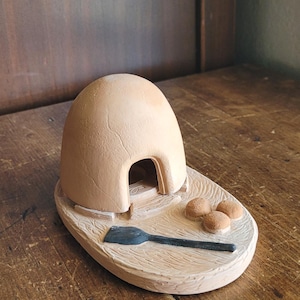 May include: A light tan, dome-shaped clay structure with an arched opening, set on an oval base. The base features a textured surface with three small, round, tan clay spheres and a single, flat, black, elongated tool placed in front of the opening.