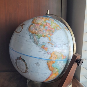 May include: A vintage globe showing a detailed world map with colourful continents and oceans. It is mounted on a wooden and metal stand. The globe has a blue meridian ring and a legend label.