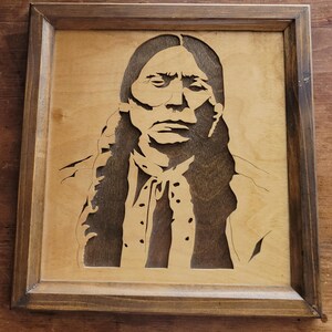 May include: Wooden framed artwork featuring a portrait of a Native American man. The image is a detailed cutout, with dark brown hair and facial features against a lighter wood background. The frame is a darker brown.