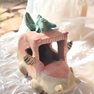 May include: A whimsical, hand-painted ceramic car sculpture in shades of pink, cream, and teal. The car features a rounded body, open windows, and decorative stripes. A teal creature sits atop the roof, adding a playful touch to this decorative piece.