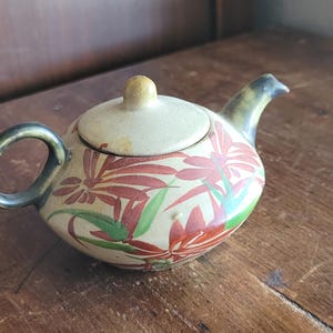 May include: A small ceramic teapot with a cream-colored base featuring a painted pattern of red flowers and green leaves. The teapot has a dark green handle and spout, and a matching cream-colored lid with a small knob handle on the top.