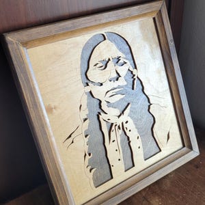 May include: Wooden framed artwork featuring a portrait of a Native American man. The portrait is laser-cut from wood, with a light-colored background and a darker, gray-toned figure. The frame is a medium brown color, adding a rustic touch to the piece.
