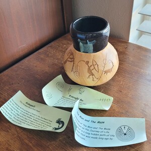 May include: A handcrafted gourd vase with a dark brown rim and etched designs of figures and symbols. The vase is accompanied by informational cards with text and illustrations. The vase is on a wooden surface.