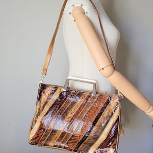 May include: A rectangular handbag featuring a glossy, multi-colored snakeskin-patterned exterior with diagonal stripes in shades of brown, tan, and black. The bag includes a metallic silver-toned top handle and a thin, adjustable tan shoulder strap.