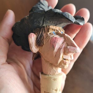 May include: Hand-carved wooden bottle stopper depicting a face with a black hat. The face has pink cheeks, blue eyes, and a large nose. The stopper is made of cork.