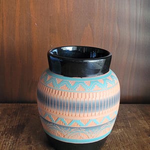 Op de afbeelding: Een handgemaakte keramische vaas met een zwarte rand en voet. De body heeft een gedetailleerd patroon in tinten perzik, teal en grijs. De vaas heeft een ronde vorm en wordt waarschijnlijk gebruikt voor decoratieve doeleinden.