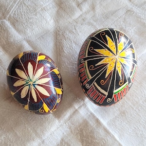 Könnte beinhalten: Zwei dekorative Eier mit aufwendigen handbemalten Mustern. Das linke Ei hat eine dunkelrote Basis mit einem cremefarbenen Blumenmuster und gelben Akzenten. Das rechte Ei hat eine schwarze Basis mit einem gelben Stern und roten Details.