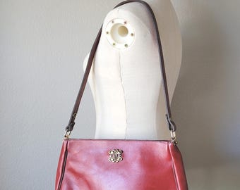May include: A red leather shoulder bag with a brown strap. The bag has a gold-colored decorative emblem on the front. The bag is displayed on a mannequin.