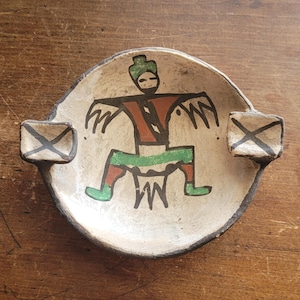May include: A round ceramic ashtray with a cream-colored base and a dark rim. The center features a hand-painted figure in red, green, and black. Two rectangular rests extend from the sides, each marked with a black cross pattern on the top surface.