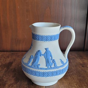 Puede incluir: Jarra de cerámica blanca con bandas decorativas azules y figuras en relieve. La jarra tiene un asa blanca y un pico. Los relieves azules representan figuras clásicas. La jarra está sobre una superficie de madera.