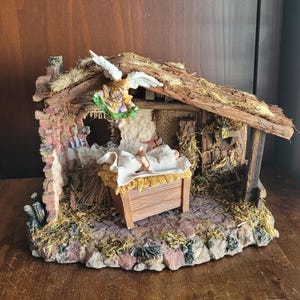 May include: A detailed nativity scene figurine. The scene features a baby in a manger, a rustic stable with a thatched roof, and an angel. The figurine is made of painted resin and has a detailed, textured appearance. The color palette includes browns, greens, and whites.