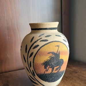 May include: A tan-coloured vase with a black rim and a design depicting a Native American on horseback against a sunset. The vase has a textured surface and black leaf-like accents.