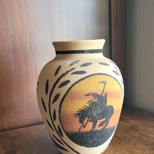 May include: A beige ceramic vase with a textured sand finish featuring a black silhouette of a person riding a bucking horse inside a circular sunset-colored gradient of yellow and orange, accented by black leaf-like patterns around the rim.