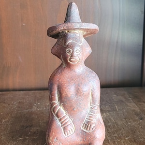May include: A reddish-brown terracotta figurine of a seated figure wearing a wide-brimmed pointed hat. The figure features incised lines on the arms and face, with a smooth, matte texture throughout the body and a distinct, rounded head shape.
