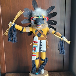 May include: A handcrafted Native American Kachina doll. The doll features a black face with a yellow body and arms. It is adorned with feathers, fur, and decorative elements, holding bundles in each hand. The doll stands on a wooden base.