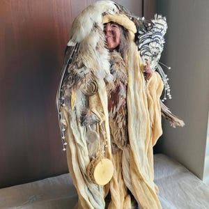 May include: A handcrafted doll dressed in a beige and brown outfit, adorned with feathers and a feathered headdress. The doll holds a bundle of feathers and a small drum. The doll's face has a serene expression.