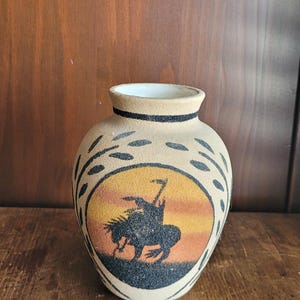 Könnte beinhalten: Eine beige Keramikvase mit schwarzem Rand und einem indianischen Design. Die Vase zeigt eine Silhouette eines Indianers auf einem Pferd vor einem Sonnenuntergang. Schwarze blattartige Muster verzieren die Seiten.