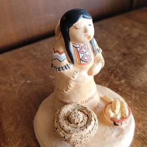 Può includere: Statuina in ceramica di una donna nativa americana in abito beige con capelli neri. La statuina ha un cestino e una ciotola con grano su una base beige. L'abito della donna presenta motivi geometrici rossi, bianchi e blu.