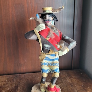 May include: A handcrafted figurine of a Native American figure. The figure wears a hat with feathers, a red sash, and blue and yellow patterned shorts. The figure is standing on a wooden base.