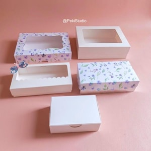 May include: A selection of white cardboard boxes, some with clear windows, and others decorated with floral patterns in purple and green. One box has a scalloped edge. The boxes are on a pink surface.