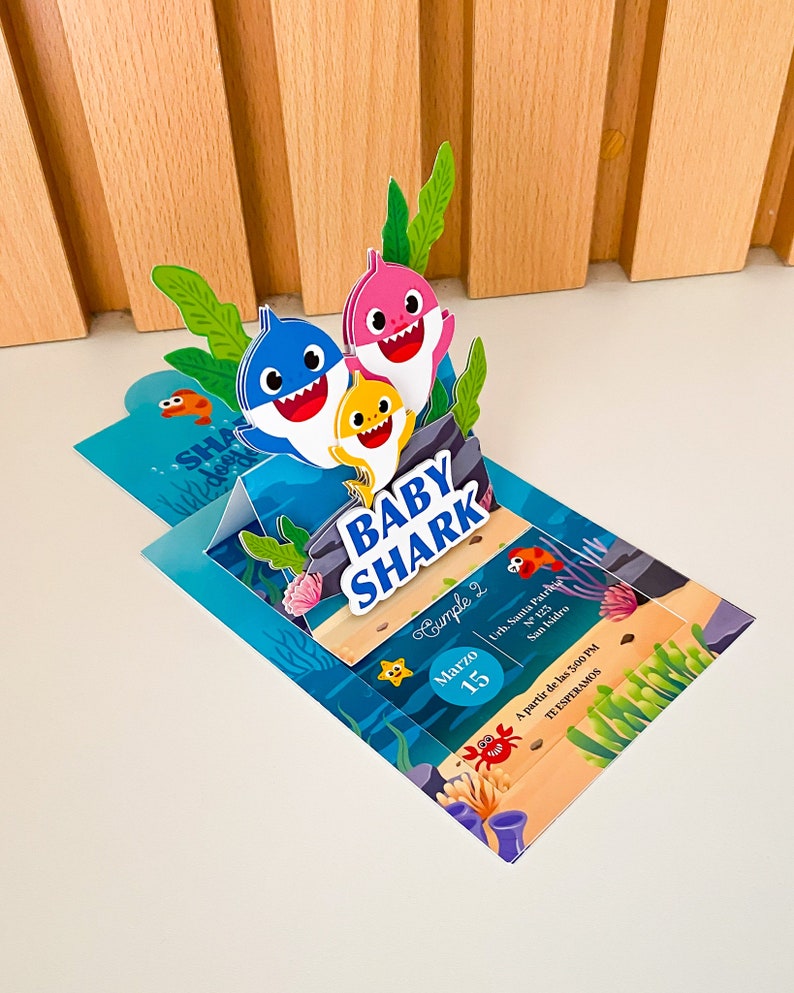 K&ouml;nnte beinhalten: Eine blaue, rosa und gelbe Baby Shark Pop-up-Karte mit Einladung. Die Karte zeigt ein 3D-Pop-up der Baby Shark Familie. Der Einladungstext lautet "Cumple 2" (Geburtstag 2), "Marzo 15" (15. M&auml;rz), "Urh. Santa Patri N 123 San Isidro" (Santa Patri Stra&szlig;e N 123 San Isidro), "A partir de las 3:00 PM" (Ab 15:00 Uhr) und "TE ESPERAMOS" (Wir erwarten Sie).